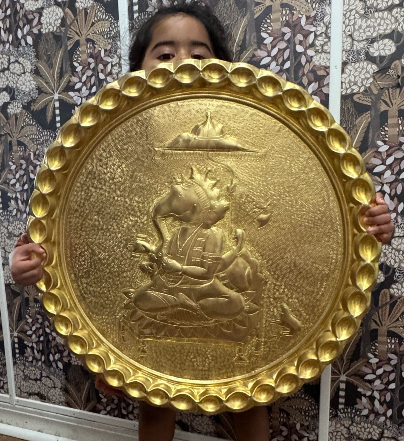 Brass decorative plate with a central figure held by a child against a patterned wall.